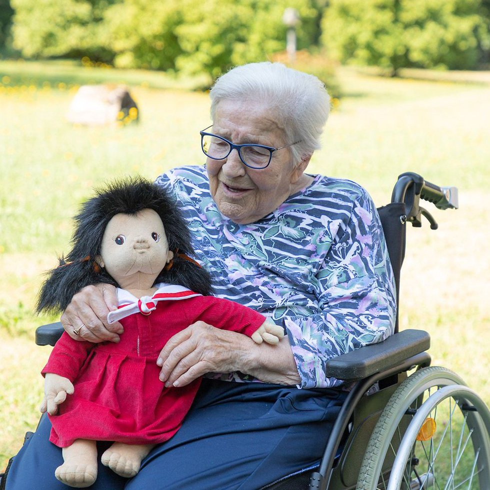 Seniorin im Rollstuhl hält die Joyk Empathiepuppe Mia im Arm