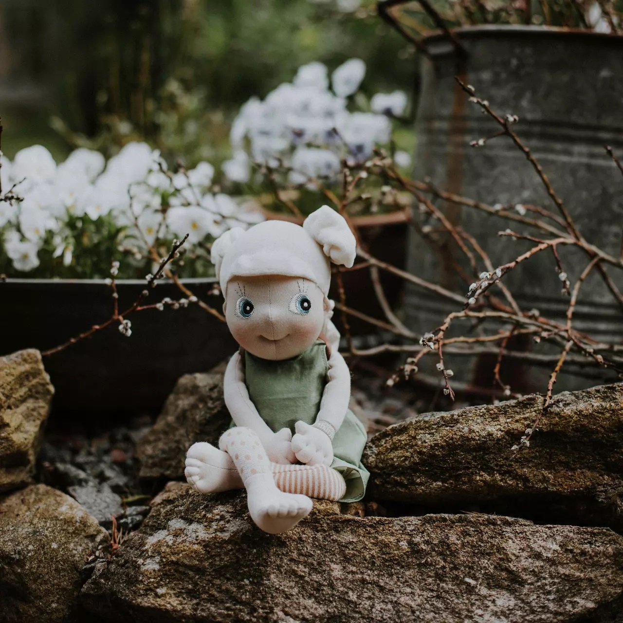 Rubens EcoBuds Puppe im grünen Frühlings-Outfit sitzt auf einer Gartenmauer