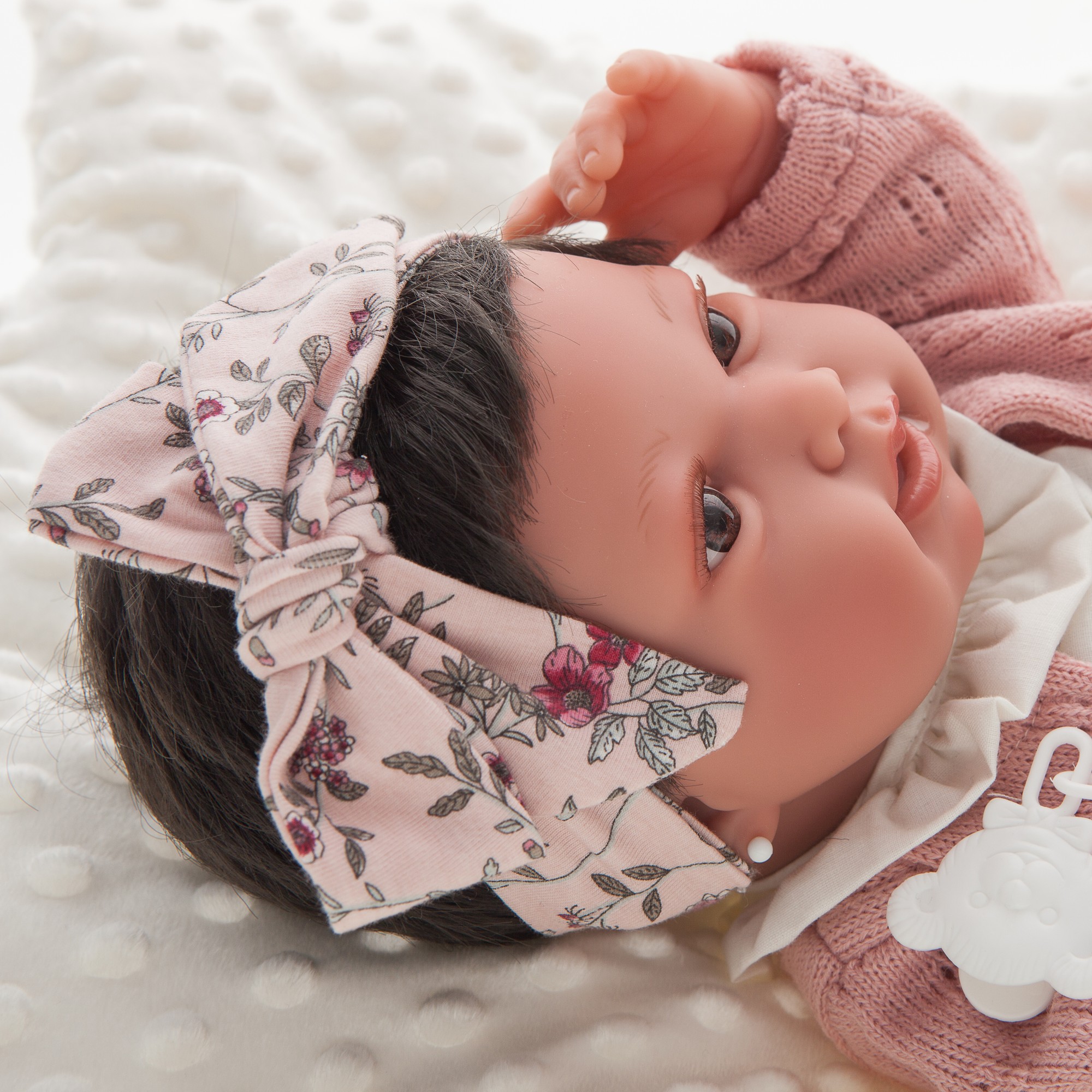 Antonio Juan baby doll Pipa, close-up of the face with dark hair, floral bow and pink knit cardigan