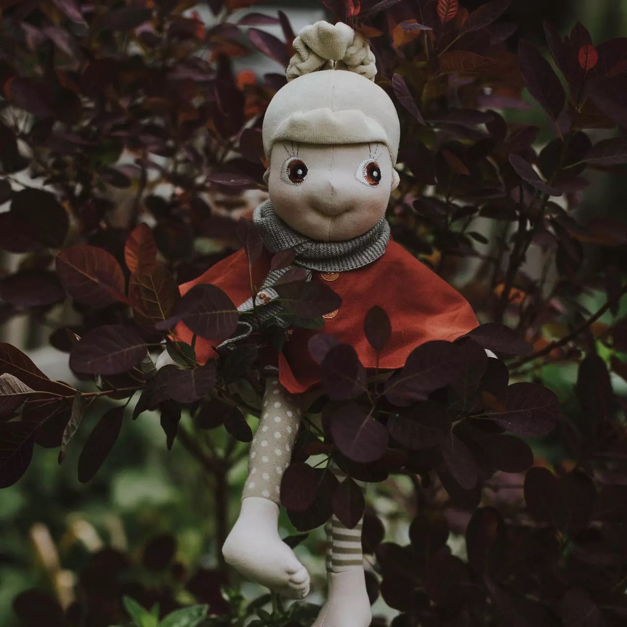 Rubens EcoBuds Puppe im rostroten Herbst-Poncho zwischen dunklem Herbstlaub
