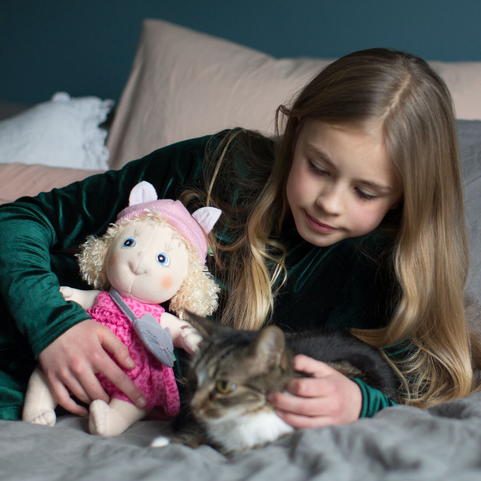 Kind mit der Rubens Cutie Puppe Emelie streichelt eine Katze auf dem Bett