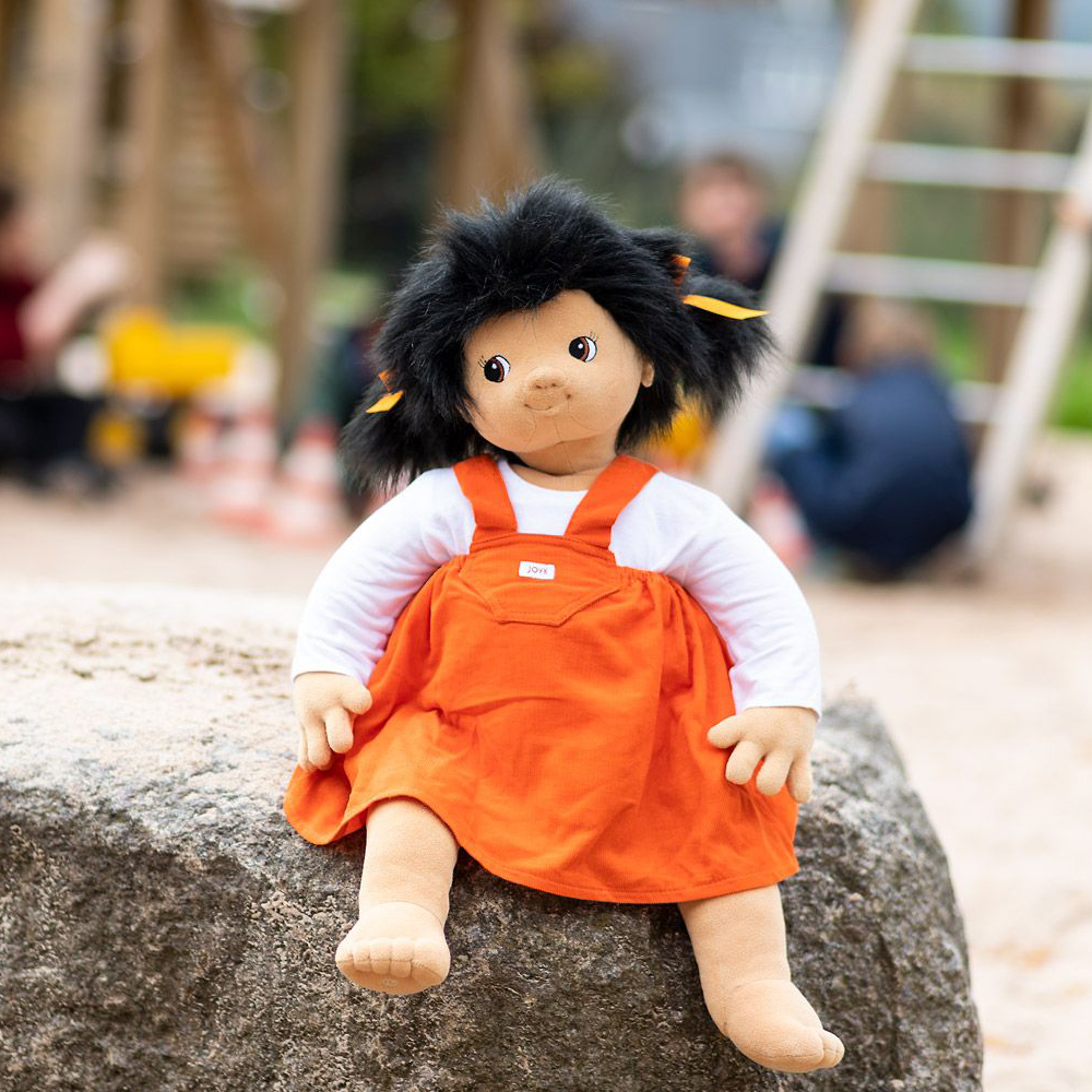Joyk Empathiepuppe Emelie sitzt auf einem Findling am Spielplatzrand
