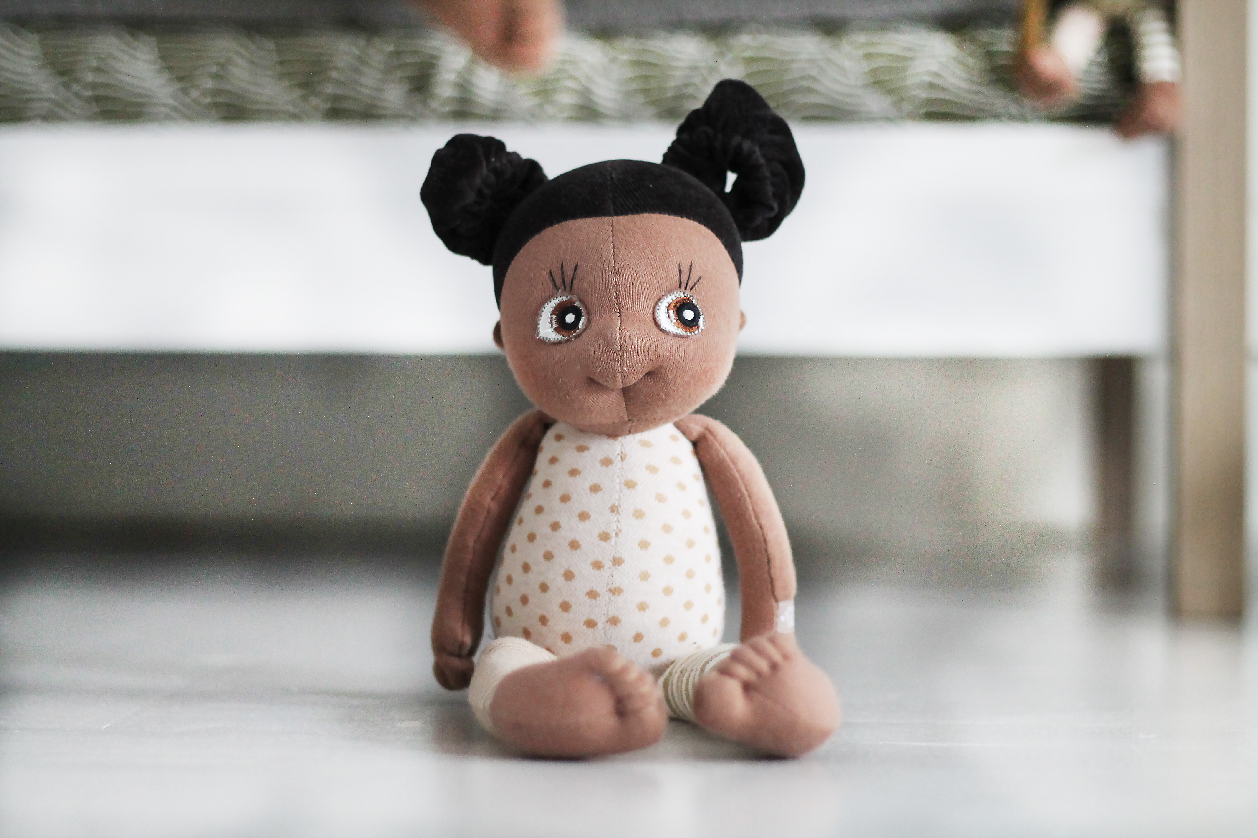 Rubens EcoBuds Mini-Puppe Flora sitzt mit gepunktetem Top vor einem Bett